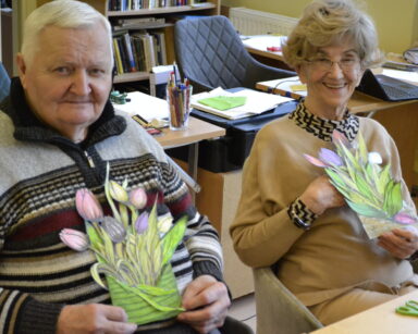 Zbliżenie. Dwoje seniorów pozuje do zdjęcia z wyciętymi i pokolorowanymi kwantami tulipanów. Seniorzy uśmiechają się.