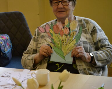 Zbliżenie. Seniorka w swetrze w kratkę siedzi na krześle i trzyma w rękach bukiet wyciętych i pomalowanych tulipanów.