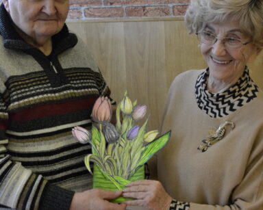 Zbliżenie. Para seniorów pozuje do zdjęcia. Para uśmiech się, w dłoniach trzyma bukiet wyciętych i pomalowanych kwiatów.