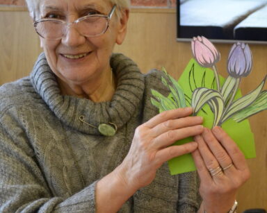 Zbliżenie. Uśmiechnięta seniorka w szarym swetrze, pozuje do zdjęcia, trzyma w dłoniach wycięte i pomalowane tulipany.