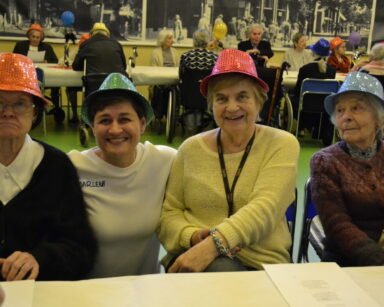 Sala. Cztery kobiety w karnawałowych kapeluszach pozują do zdjęcia. W tle seniorzy przy stołach.