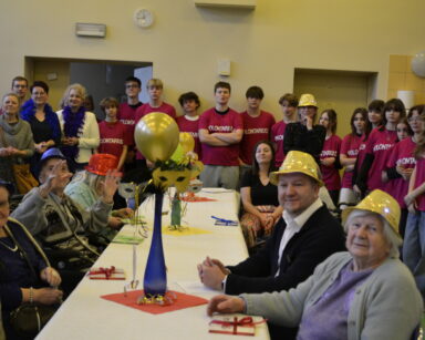 Sala. Grupa wolontariuszy, seniorów i opiekunów pozuje do zdjęcia. Seniorzy siedzą przy stole w karnawałowych kapeluszach.