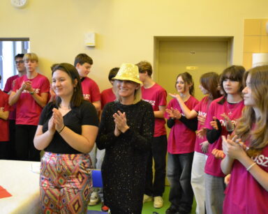 Sala. Dwie kobiety i kilkunastu wolontariuszy w różowych podkoszulkach klaszczą w ręce.