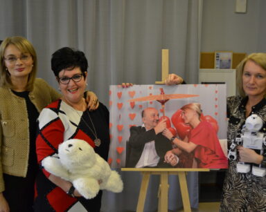 Trzy kobiety pozują do zdjęcia przy fotografii pary seniorów. Dwie kobiety trzymają fokę Paro i robota JD.