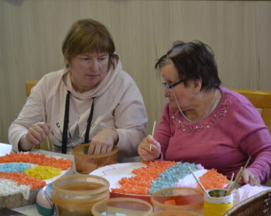Dwie seniorki siedzą przy stole. Na stole styropianowe jajko ozdabiane kolorowymi materiałami przez 2 kobiety.