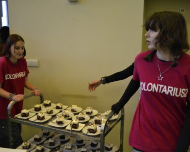 Zbliżenie. Dwie wolontariuszki ciągną wózek z pokrojonymi kawałkami czekoladowego ciasta.