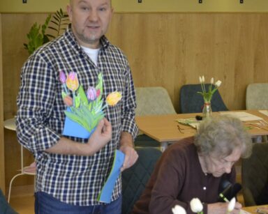 Sala. Mężczyzna trzyma w dłoni wycięte kwiaty. Obok siedzi przy stole seniorka i koloruje tulipany.