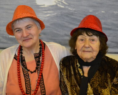 Zbliżenie. Dwie seniorki w cekinowych, karnawałowych kapeluszach pozują do zdjęcia. Kobiety uśmiechają się.