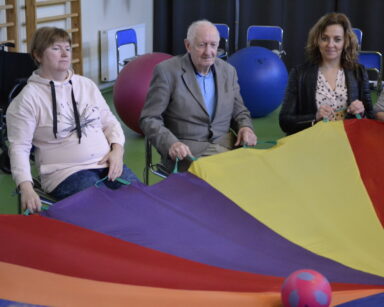 Zbliżenie. Cztery osoby siedzą na krzesłach. W rękach trzymają kolorowa płachtę, a na niej mała piłka.