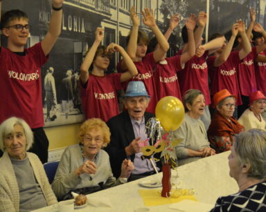 Sala. Grupa seniorów siedzi przy stole. Obok wolontariusze. Wszyscy unoszą ręce do góry i uśmiechają się.