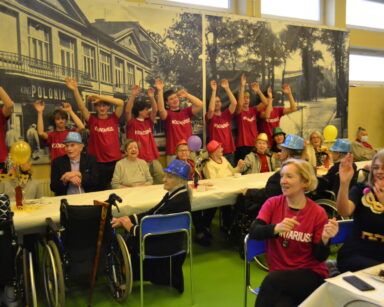 Sala. Grupa seniorów przy stolach i wolontariuszy przy ścianie unosi ręce do góry. Wszyscy śpiewają. W tle zdjęcia.