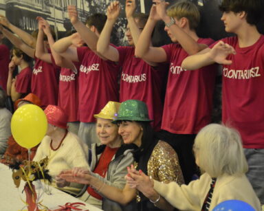 Sala. Grupa seniorów przy stolach i wolontariuszy przy ścianie unosi ręce do góry. Wszyscy śpiewają.