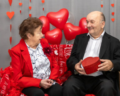 Zbliżenie. Para seniorów pozuje do zdjęcia. Senior trzyma pudełko w kształcie serca. W tle balony i ściana serc.