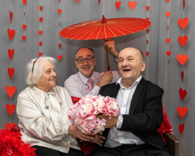 Zbliżenie. Para seniorów uśmiecha się i trzyma bukiet kwiatów. Nad parą mężczyzna trzyma czerwony japoński parasol.