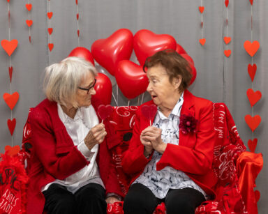 Zbliżenie. Dwie uśmiechnięte seniorki trzymają w ręku lizaki serca. W tle balony w serca i ściana serduszek.