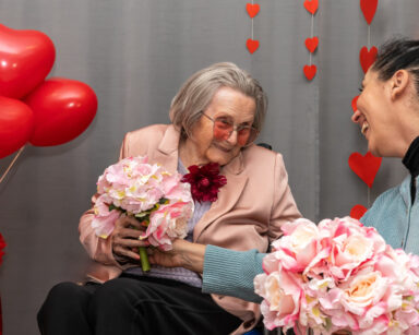 Zbliżenie. Kobieta podaje uśmiechniętej seniorce kwiaty. W tle balony serca i na ścianie girlandy z serc.