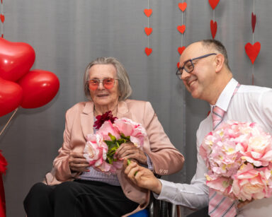 Zbliżenie. Mężczyzna klęczy i podaje uśmiechniętej seniorce bukiet kwiatów. Seniorka ma na sobie różowe okulary.