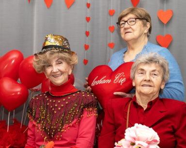Trzy seniorki pozują do zdjęcia. Kobiety ubrane są w czerwień. Jedna z pań trzyma poduszkę serce z napisem 
