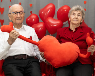 Para seniorów pozuje do zdjęci. W dłoniach trzymają serce z rękoma. W tle czerwone balony i serduszka.