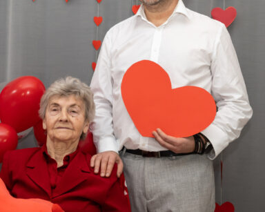 Para pozuje do zdjęcia. Mężczyzna stoi obok siedzi seniorka. Kobieta trzyma w ręku pluszowe serce. W tle balony, serca.