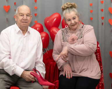 Para pozuje do zdjęcia. Kobieta wskazuje na pantofel, który trzyma mężczyzna. W tle czerwone balony, serca.