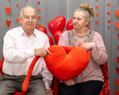 Zbliżenie. Para pozuje do zdjęcia. Seniorzy trzymają pluszowe serce. W tle czerwone balony, serca.