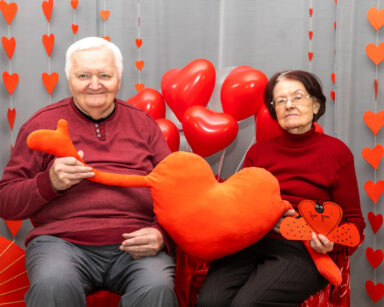 Zbliżenie. Para seniorów trzyma w ręku serce z rączkami. Seniorzy pozują do zdjęcia. W tle balony, serca.