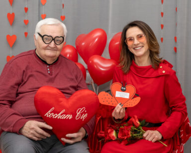 Zbliżenie. Para pozuje do zdjęcia. Senior na nosie ma okulary w serca, w ręku poduszka serce. Obok kobieta trzyma róże.