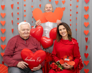 Zbliżenie. Trzy osoby pozują do zdjęcia. Trzymają w dłoniach serca. Kobieta trzyma róże i kartkę serce.