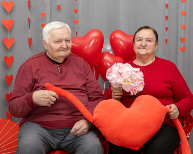 Zbliżenie. Para pozuje do zdjęcia. Ubrani są na czerwono, w dłoniach trzymają serce i kwiaty. W tle balony i serca.