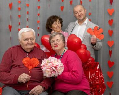 Zbliżenie. Dwie pary pozują do zdjęcia. W dłoniach trzymają serca, kwiaty. W tle czerwone balony i serduszka.