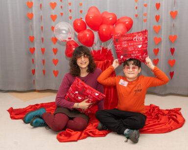 Para dzieci pozuje do walentynkowego zdjęcia na czerwonym tle balonów, koca i poduszek.