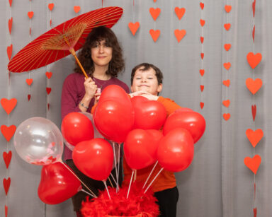 Para dzieci pozuje do zdjęcia. Dziewczynka trzyma czerwony parasol, chłopczyk opiera się o ozdobę z balonów.