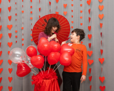 Para dzieci próbuje ugryźć lizaka. Dziewczynka w dłoni trzyma czerwony parasol. Obok ozdoba z balonów i ściana serc.