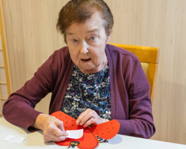 Zbliżenie. Seniorka pozuje do zdjęcia. Seidzi przy stole i klei mały liścik do wnętrza kartki serduszko, biedronka.