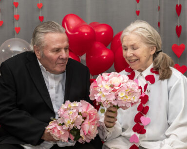 Zbliżenie. Para seniorów patrzy na kwiaty. W tle balony w serca, ściana w serca.