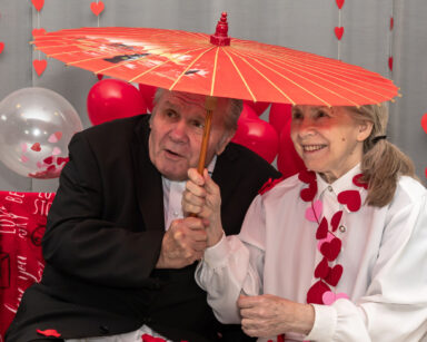 Zbliżenie. Uśmiechnięta para seniorów siedzi pod czerwoną parasolką. W tle balony i ściana w czerwone serca.
