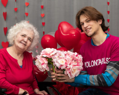 Zbliżenie. Młody wolontariusz przekazuje kwiaty zamyślonej seniorce. Kobieta ubrana jest w czerwień. W tle balony i serca.