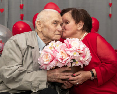 Zbliżenie. Seniorka daje buziaka seniorowi. Senior trzyma bukiet kwiatów. W tle balony i serca.