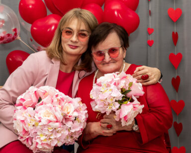 Zbliżenie. Dwie kobiety pozują do zdjęcia. Kobiety ubrane są w róż i czerwień. W rękach trzymają kwiaty.