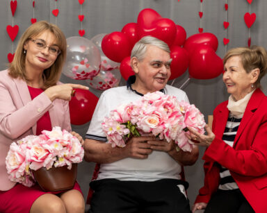 Zbliżenie. Trzy osoby pozują do zdjęcia. Jedna kobieta wskazuje na parę seniorów. Kobieta w drugim ręku trzyma kwiaty.
