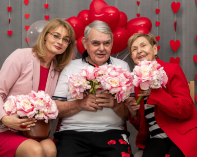 Zbliżenie. Trzy osoby z kwiatami w rękach pozują do zdjęcia. Kobiety ubrane są w róż, mężczyzna w biel. W tle ściana serc.
