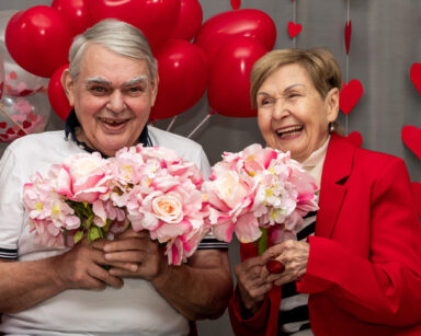 Para seniorów pozuje do zdjęcia. Oboje w dłoniach trzymają kwiaty i uśmiechają się. W tle czerwone balony i serca.