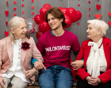 Młody wolontariusz trzyma od ręce dwie uśmiechnięte seniorki. Kobiety ubrane są w róż. W tle balony, serduszka na ścianie.