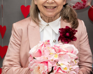 Zbliżenie. Uśmiechnięta seniorka pozuj do zdjęcia. Ubrana jest w różowy garnitur. W dłoniach trzyma bukiet kwiatów.
