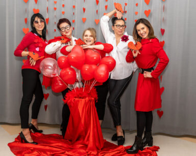 Grupa kobiet pozuje do sesji walentynkowej. Przed kobietami stoi ozdoba z czerwonych balonów. W tle ściana w serca.