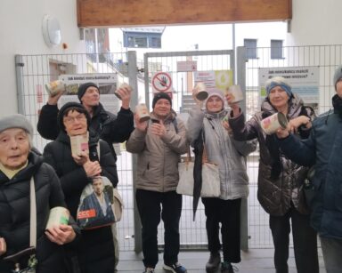 Grupa osób pozuje do zdjęcia przy wejściu do schroniska. W rękach trzymają karmę dla zwierząt.