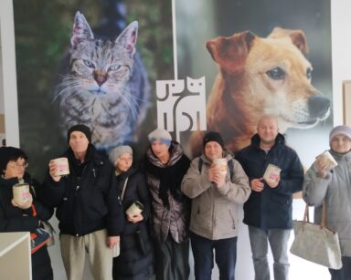 Sala. Grupa osób z puszką karmy w rękach pozuje do zdjęcia pod fotografiami kota i psa.