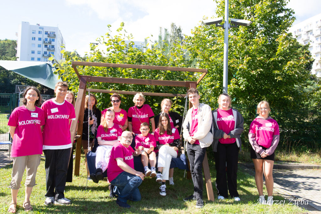 Grupa młodych wolontariuszy z seniorem wolontariuszem w różowych podkoszulkach pozują do zdjęcia grupowego.