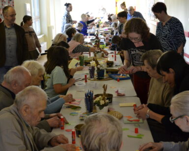 Sala. Długi stół. Przy stole seniorzy i dzieci wyklejają ozdoby na świąteczne kartki wielkanocne.